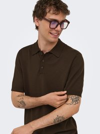 Jeune homme portant un polo marron foncé à manches courtes, des lunettes, montrant des tatouages de scorpion, boussole et crabe sur ses avant-bras.