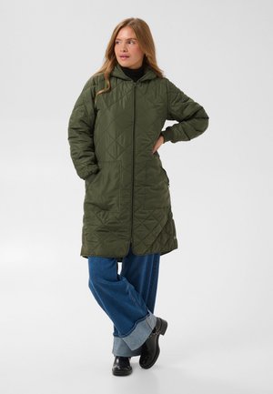 Une femme porte un long manteau matelassé vert olive à capuche, un jean bleu retroussé et des bottes noires sur un fond uni.