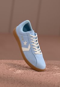 Lyseblå semsket sneaker med hvite detaljer, som har en stjerne logo, hvite snører og en brun gummisåle. Glatt tekstur, lav profil design.