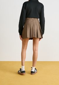 Gonna a pieghe marrone esposta con un top nero, abbinata a scarpe da ginnastica nere, calzini bianchi e un pavimento giallo senape.