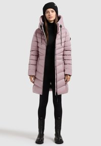 Kvinde iført en lyse mauve pufferjakke med hætte, sort beanie, sorte leggings og sorte ankelstøvler, stående mod en ensfarvet baggrund.