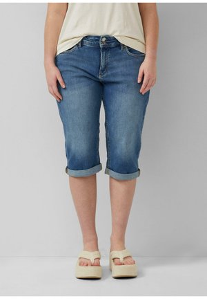 Osoba nosząca niebieskie dżinsowe spodnie capri podwinięte na nogawkach i kremowe sandały na platformie, stojąca na jednolitym szarym tle.