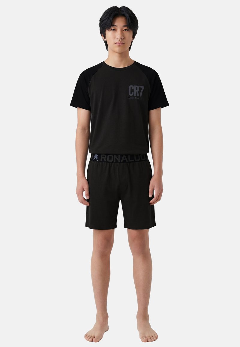 Jeune homme debout pieds nus portant un t-shirt noir CR7 Nightwear à manches courtes et un short assorti avec une ceinture marquée Ronaldo.