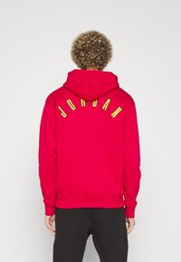 Sudadera con capucha roja que presenta letras amarillas de "JORDAN" en la parte posterior. El material parece suave, con un ajuste relajado y puños acanalados.