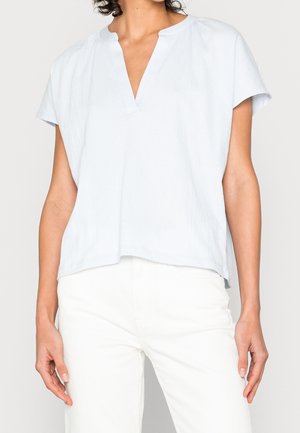Vrouw met een losse lichtblauwe blouse met korte mouwen en V-hals en een witte broek, afgebeeld van schouders tot dijen tegen een effen achtergrond.