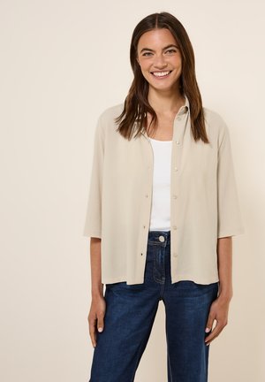 Camicia button-up beige con vestibilità rilassata, maniche corte e tessuto testurizzato, abbinata a jeans di denim blu scuro.