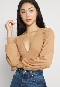 Camisola comprida bege, em tecido canelado, com um decote em V profundo e mangas bufantes, combinada com calças de ganga de cintura alta. Tecido texturizado visível.