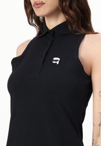 Polo negro sin mangas con collar, con una tapeta frontal y un pequeño logotipo blanco en el lado izquierdo del pecho. Tejido texturizado.