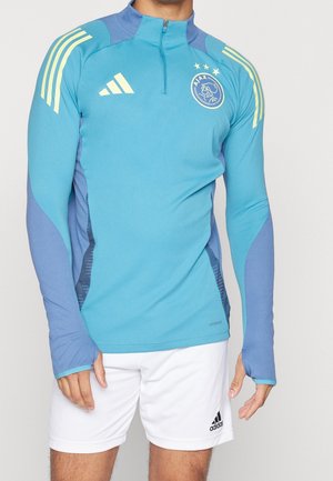 Hombre con sudadera de entrenamiento azul de Ajax con cremallera hasta el pecho, con rayas amarillas, y pantalones cortos blancos de Adidas, sobre un fondo liso.