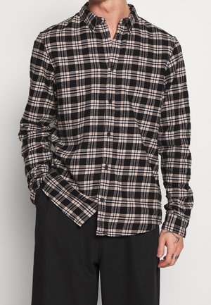 Uomo che indossa una camicia a quadri nera, bianca e beige con maniche lunghe e bottoni, infilata nei pantaloni neri, con una mano in tasca e tatuaggi visibili sulla mano.