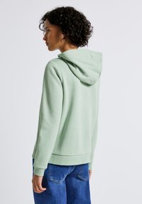 Felpa con cappuccio verde chiaro, dalla vestibilità comoda, con polsini e orlo in rilievo. Tessuto morbido, indossata con pantaloni in denim blu.