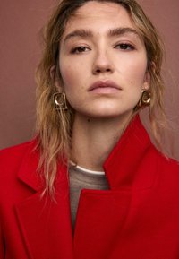 Rød uldfrakke med en struktureret krave, stylet over en beige top. Guld firkantede øreringe tilføjer en metallisk accent. Baggrunden er varm brun.