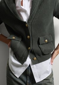 Cardigan lavorato a maglia verde scuro con bottoni dorati e tasche, indossato sopra una camicia bianca con bottoni. Tessuto testurizzato con design a coste.