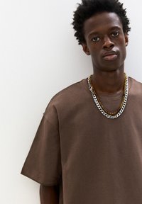 Braunes, übergroßes T-Shirt aus weichem Material mit rundem Halsausschnitt. Kombiniert mit dreilagigen Kettenhalsschmuck in unterschiedlichen Farben und Stilen.