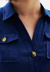 Camicia in lino blu navy con scollo a V, maniche corte, due tasche sul petto e bottoni rotondi con una leggera lucentezza.