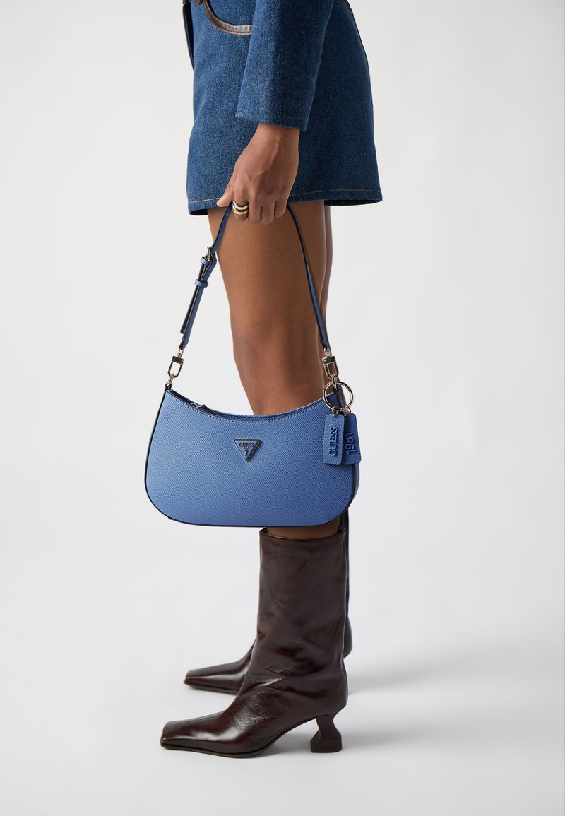 Mala texturizada azul com um logótipo triangular, alça curta e uma etiqueta, combinada com botas castanhas até ao joelho. Forma simples e curva.