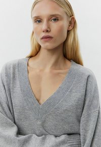 Pulover gri cu decolteu în V, textură ribbată, mâneci largi și croială lejeră, evidențiind un material moale și un design minimalist.