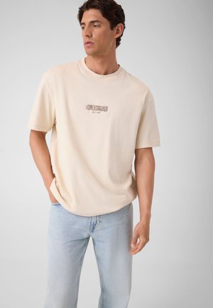 Man draagt een beige Guess T-shirt en lichtblauwe jeans, staat met één hand in zijn zak, kijkt naar links tegen een effen achtergrond.