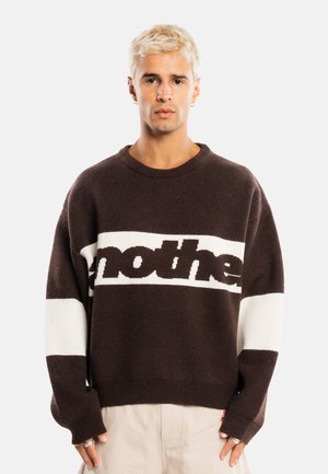Jeune personne aux cheveux blonds courts portant un pull marron foncé avec des rayures blanches horizontales et un pantalon beige sur fond uni.