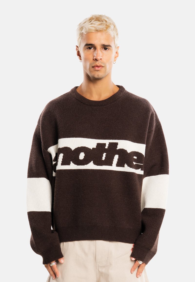 Jeune personne aux cheveux blonds courts portant un pull marron foncé avec des rayures blanches horizontales et un pantalon beige sur fond uni.