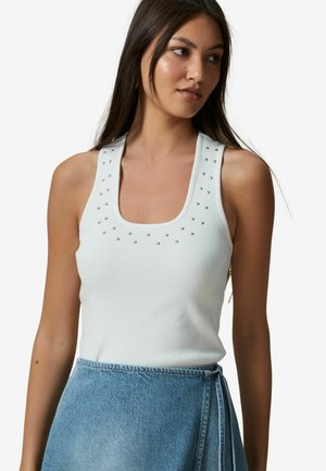 Top a righe bianco con scollatura quadrata, caratterizzato da borchie decorative argentate lungo le spalle. Indossato con una gonna di jeans azzurro chiaro.