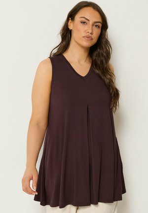 Kobieta z długimi brązowymi włosami, ubrana w ciemnobrązowy, bezrękawnikowy, luźny top z dekoltem w serek i jasne spodnie, stojąca na jednolitym tle.