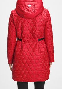 Rød quiltet jakke med hætte, prydet med et bladmønster, med bælte i taljen og et logo-patch ved halsen. Glat tekstur, midt på låret længde.