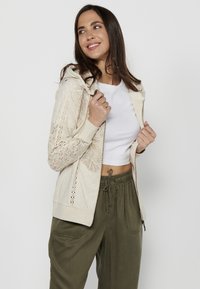 Svijetlosmeđa zip-up dukserica s cvjetnim čipkastim detaljima na rukavima, u kombinaciji s maslinasto zelenim hlačama na vezanje. Glatka tkanina, ležeran dizajn.