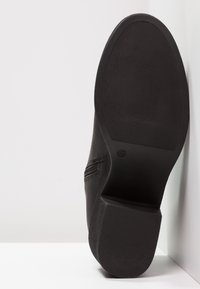 Bottine noire à cheville avec semelle en caoutchouc texturée et talon bloc, taille 42, positionnée avec la semelle vers le haut sur un fond blanc en angle.
