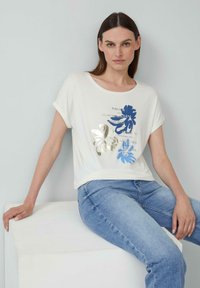 Witte t-shirt met korte mouwen, versierd met blauwe en metallic gouden bloem motieven en tekst, gecombineerd met lichtblauwe jeans, zittend op een wit platform.