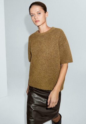 Maglione lavorato a maglia marrone e peloso con maniche corte abbinato a una gonna a matita in pelle nera. Lo scenario presenta uno sfondo grigio minimalista.