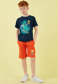 Garçon aux cheveux roux portant un t-shirt marine avec un dinosaure surfeur et un short orange avec des motifs de soleil, debout les mains dans les poches.
