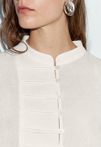 Weißes, strukturiertes Blusenshirt mit Stehkragen, ausgestattet mit fünf Knopfverschlüssen und horizontalen Falten entlang der Vorderseite. Auffällige silberne Ohrringe sind sichtbar.