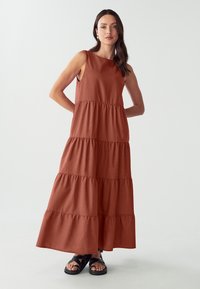Rostfarbenes Maxikleid mit ärmellosem, gestuftem Design. Hergestellt aus weichem Stoff mit glatter Textur. Kombiniert mit schwarzen Riemchensandalen.