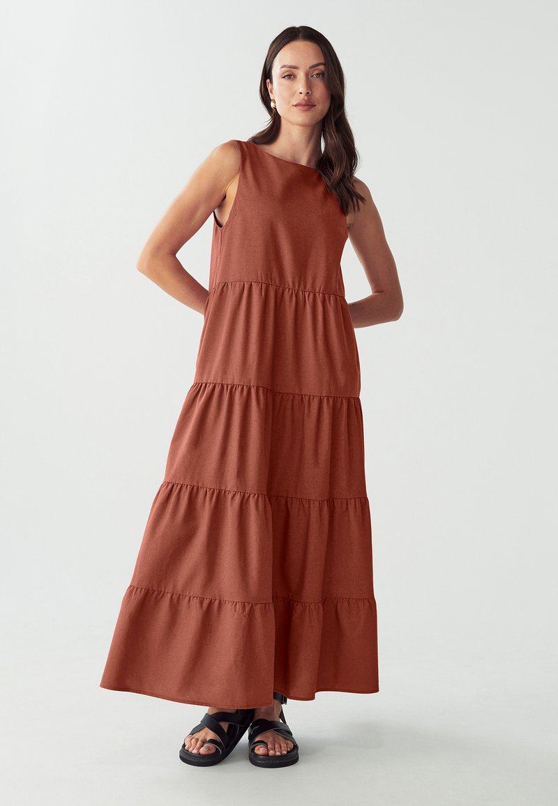 Rostfarbenes Maxikleid mit ärmellosem, gestuftem Design. Hergestellt aus weichem Stoff mit glatter Textur. Kombiniert mit schwarzen Riemchensandalen.