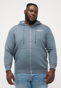 Haut à capuche zippé gris en tissu doux, avec une poche kangourou, un cordon de serrage à la capuche et un logo blanc sur la poitrine, porté avec un jean bleu.