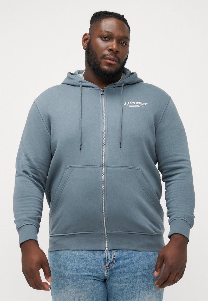 Haut à capuche zippé gris en tissu doux, avec une poche kangourou, un cordon de serrage à la capuche et un logo blanc sur la poitrine, porté avec un jean bleu.