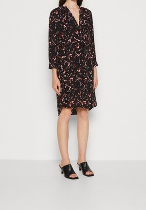 Robe noire à motifs floraux dans des nuances de rose et de violet, avec des manches longues, un décolleté en V profond et une taille ceinturée. Portée avec des mules noires.