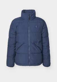 Marineblå puffjakke med høy krage, dobbel glidelås, frontlommer og en liten logo på brystet. Har et quiltet design.