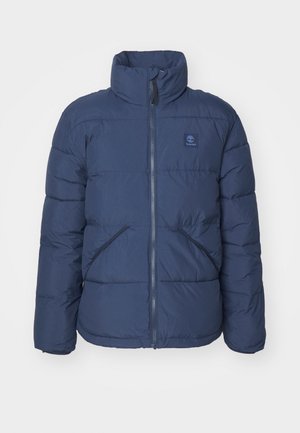 Tumši zila pūkainā džempera ar augstu apkakli, divām rāvējslēdzēja aizdara, priekšējām kabatām un mazu logotipu uz krūtīm. Iezīmējas ar kvadrātveida dizainu.