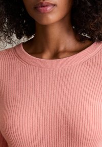 Nahaufnahme einer Person, die einen rosa gerippten Pullover trägt, mit sichtbarem Hals, Schlüsselbein und lockigem, dunklem Haar.