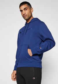 Hoodie bleu marine avec une poche avant, cordon de serrage et logo brodé. Matière en coton avec des poignets côtelés, associé à un jogging noir.
