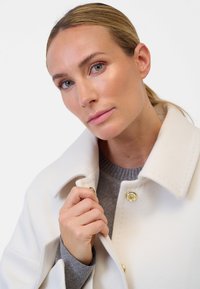 Weißer Wollmantel mit goldenen Druckknöpfen, hohem Kragen und Nahtdetails, über einem grauen Strickpullover geschichtet. Nahaufnahme des Ausschnitts und der Hand.