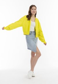 Giacca bomber gialla brillante, design corto, con polsini elasticizzati. Abbinata a una gonna in denim azzurro chiaro con uno spacco alto e orlo sfrangiato.