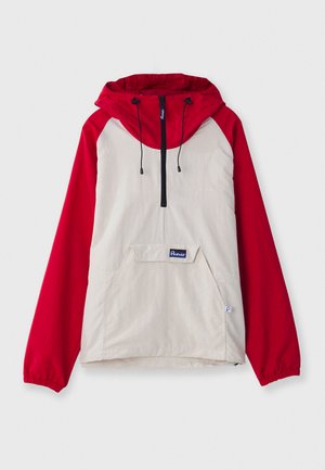 Anorakki takki, jossa on beige runko punaisilla raglan-hihansuilla, etutasku ja säädettävät nyörit hupussa. Musta vetoketjuyksityiskohta.