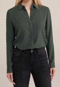 Mörkgrön skjorta med knappar och texturerat blommönster, långa ärmar och krage, i kombination med högmidjade svarta jeans.