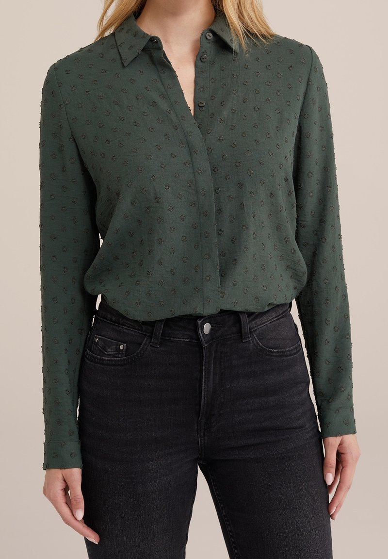 Mörkgrön skjorta med knappar och texturerat blommönster, långa ärmar och krage, i kombination med högmidjade svarta jeans.