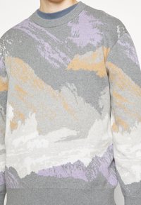 Harmaa neule, jossa on monivärinen abstrakti kuvio violetti, valkoinen ja oranssi. Ominaisuuksiin kuuluu pyöreä pääntie sekä resorit hihansuissa ja helmassa.