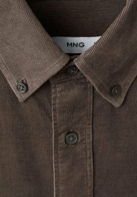 Camisa de pana marrón con cuello abotonado, presenta botones negros y tejido texturizado. La etiqueta muestra el nombre de la marca "MNG."