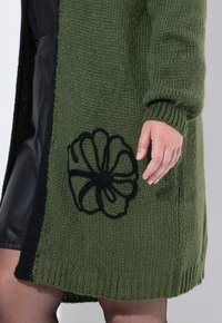 Strickjacke in Dunkelgrün mit schwarzen Akzenten, versehen mit einer großen schwarzen Blumenstickerei auf der unteren Vorderseite. Strukturiertes Material und offenes Design.